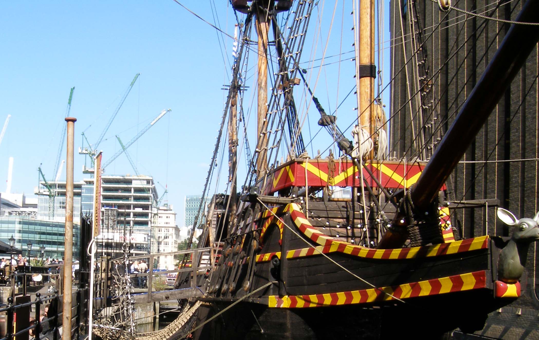 The Golden Hinde's dark wooden bow, red and yellow trim, masts and rigging at its Southwark dock, with visitors alongside.